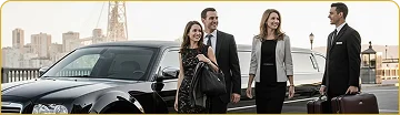 A uniformed chauffeur holding a suitcase stands next to a smiling, formally dressed couple and a female business executive as they walk beside a black Chrysler 300 stretch limousine, with a cityscape and bridge in the background.